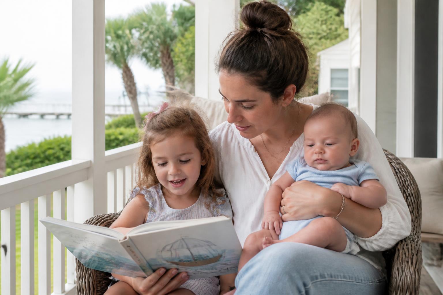 Nanny reading a picture book with a young girl while holding an infant on the porch of a Ponte Vedra home